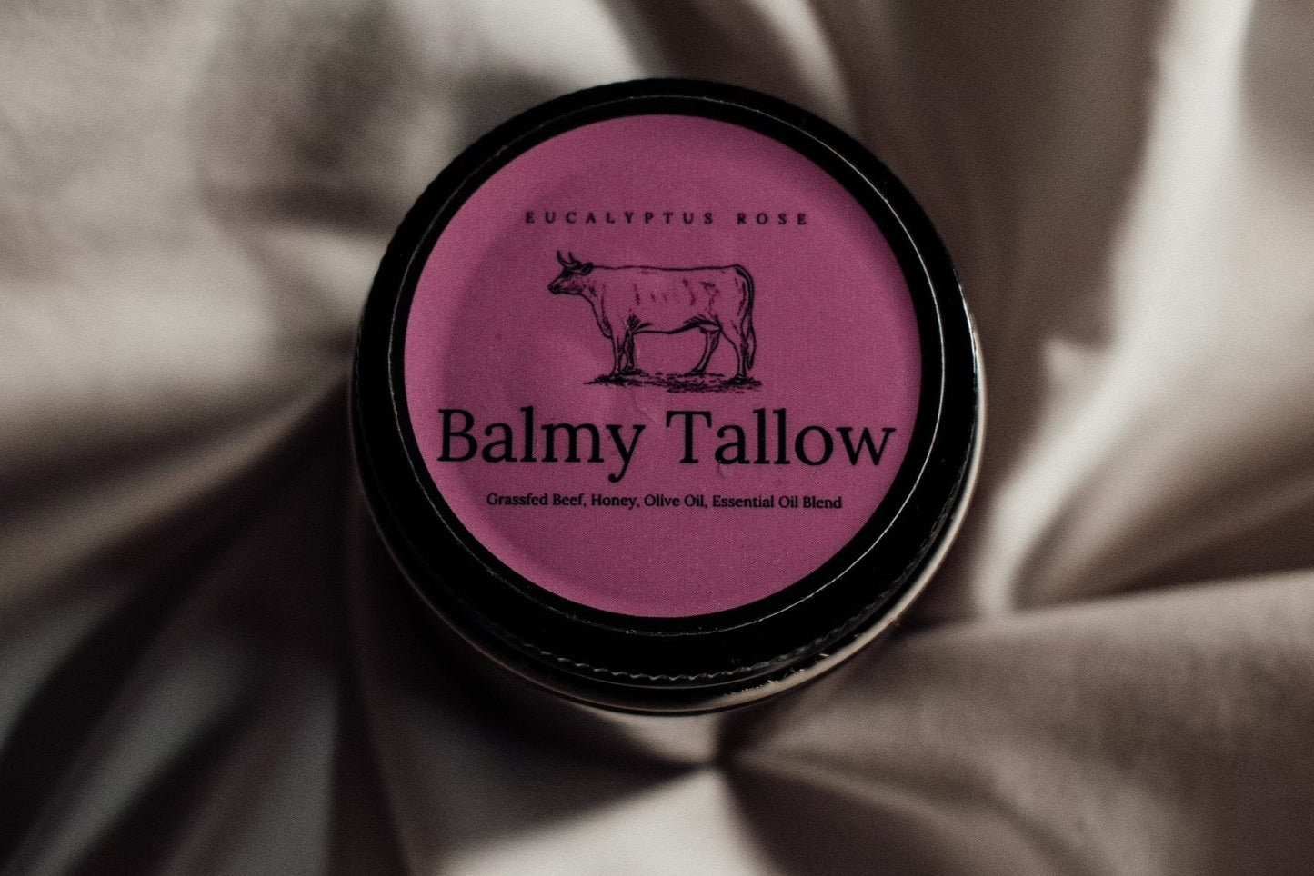 A jar of Eucalyptus Rose Tallow cream by Balmy Tallow placed on a silky fabric, with the brand name and eucalyptus leaves logo visible on the lid.