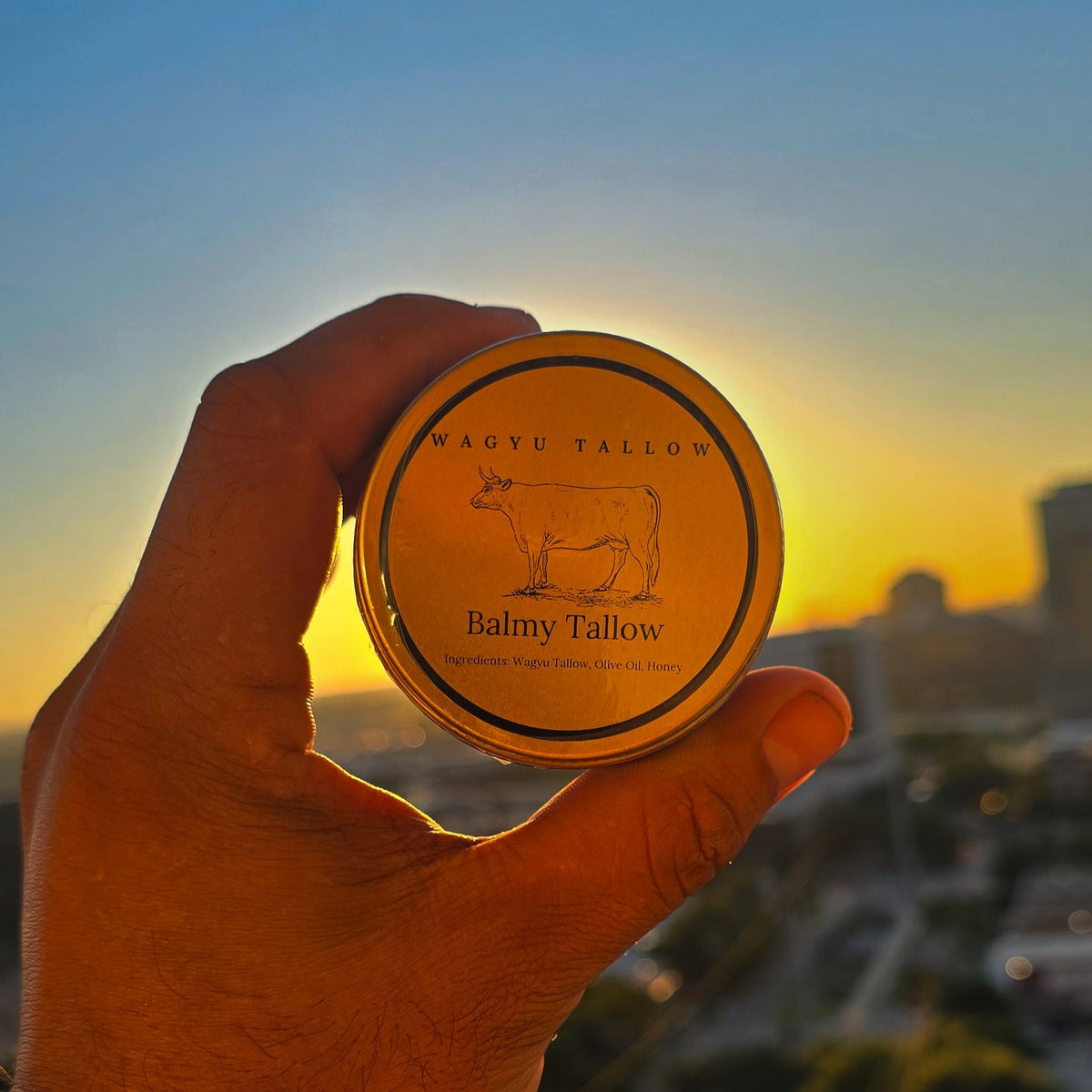 A hand holding a luxurious container labeled of Wagyu Tallow Balm in front of  cityscape background and a sunset