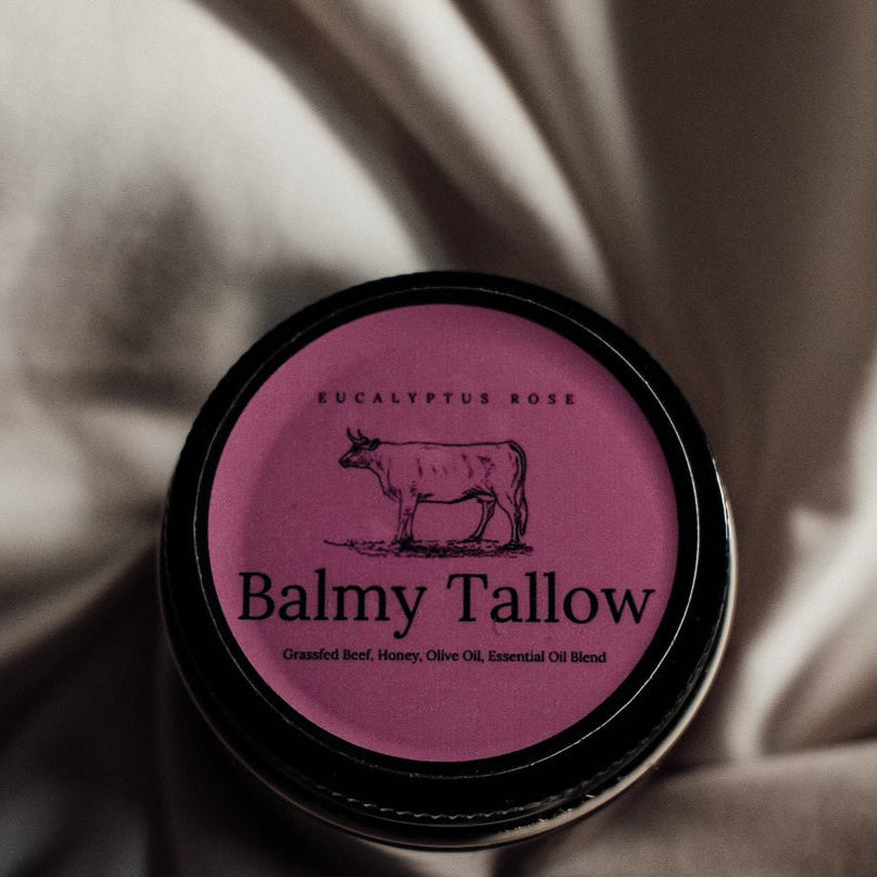 A round black jar lid with a pink label reading 'Eucalyptus Rose' and 'Balmy Tallow' with a centered cow illustration, placed on draped satin fabric.