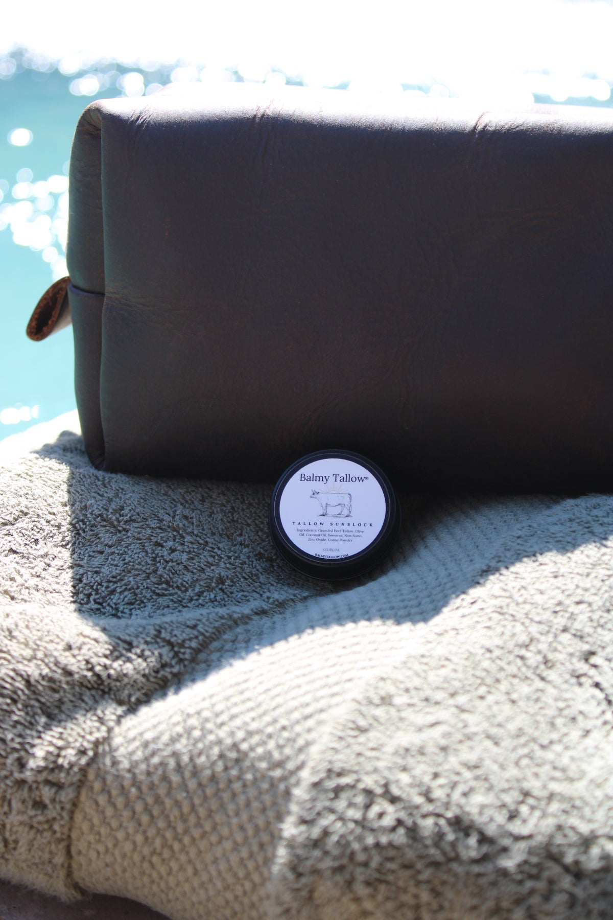 A small round tin of Balmy Tallow Sunblock sits on a textured beige towel in front of a dark brown leather pouch, with a bright outdoor water background.