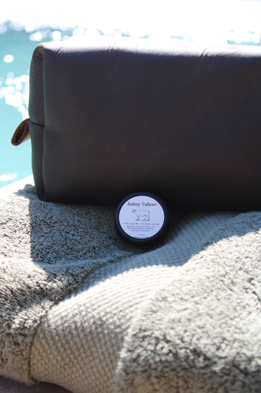 A small round tin of Balmy Tallow Sunblock sits on a textured beige towel in front of a dark brown leather pouch, with a bright outdoor water background.
