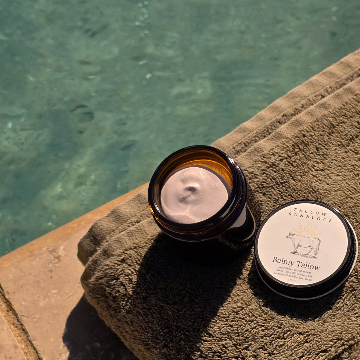 A small and opened amber glass jar filled with Balmy Tallow Sunblock with its lid placed beside it atop a folded olive green towel, set next to a swimming pool.