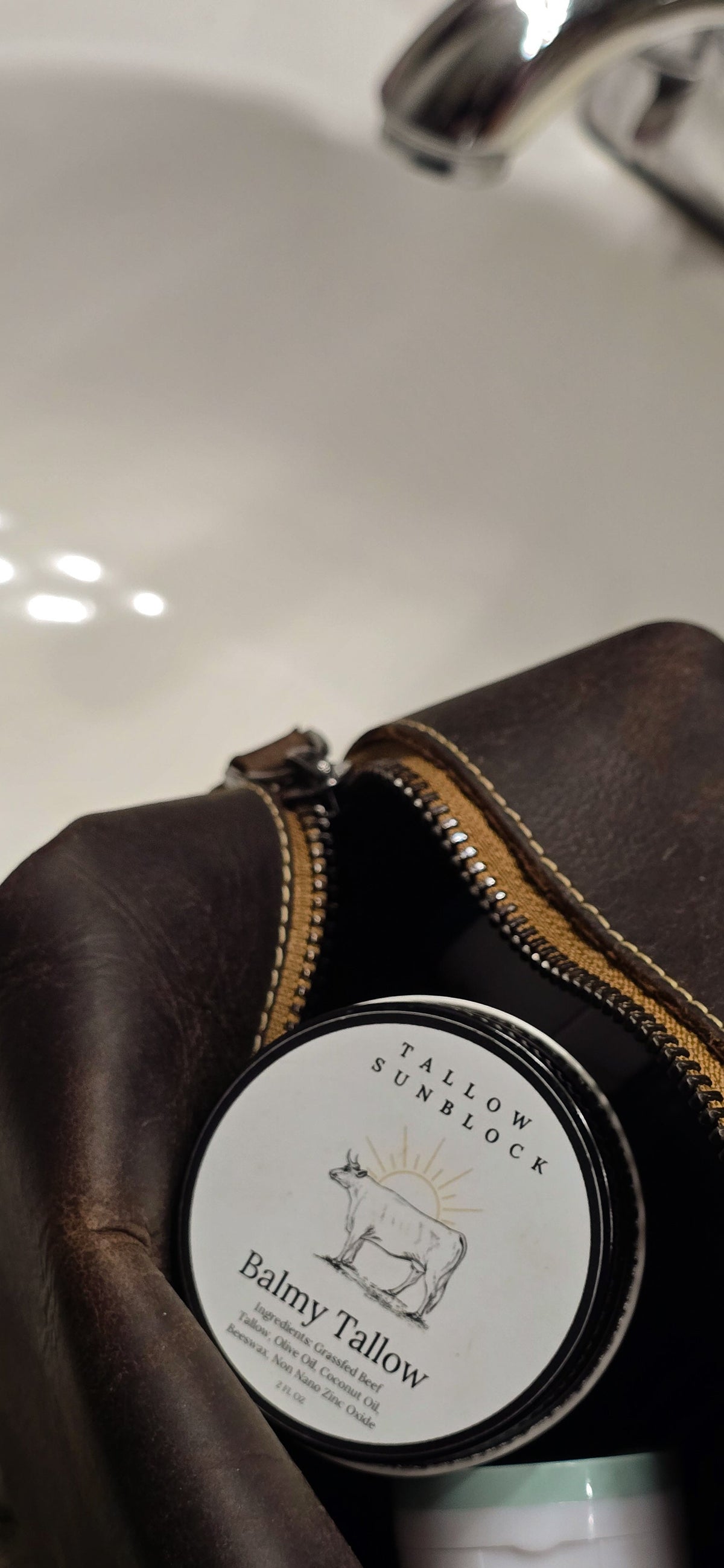 A close-up of a brown leather shower kit with Balmy Tallow's Sunblock sticking out near sink with a faucet visible in the background.