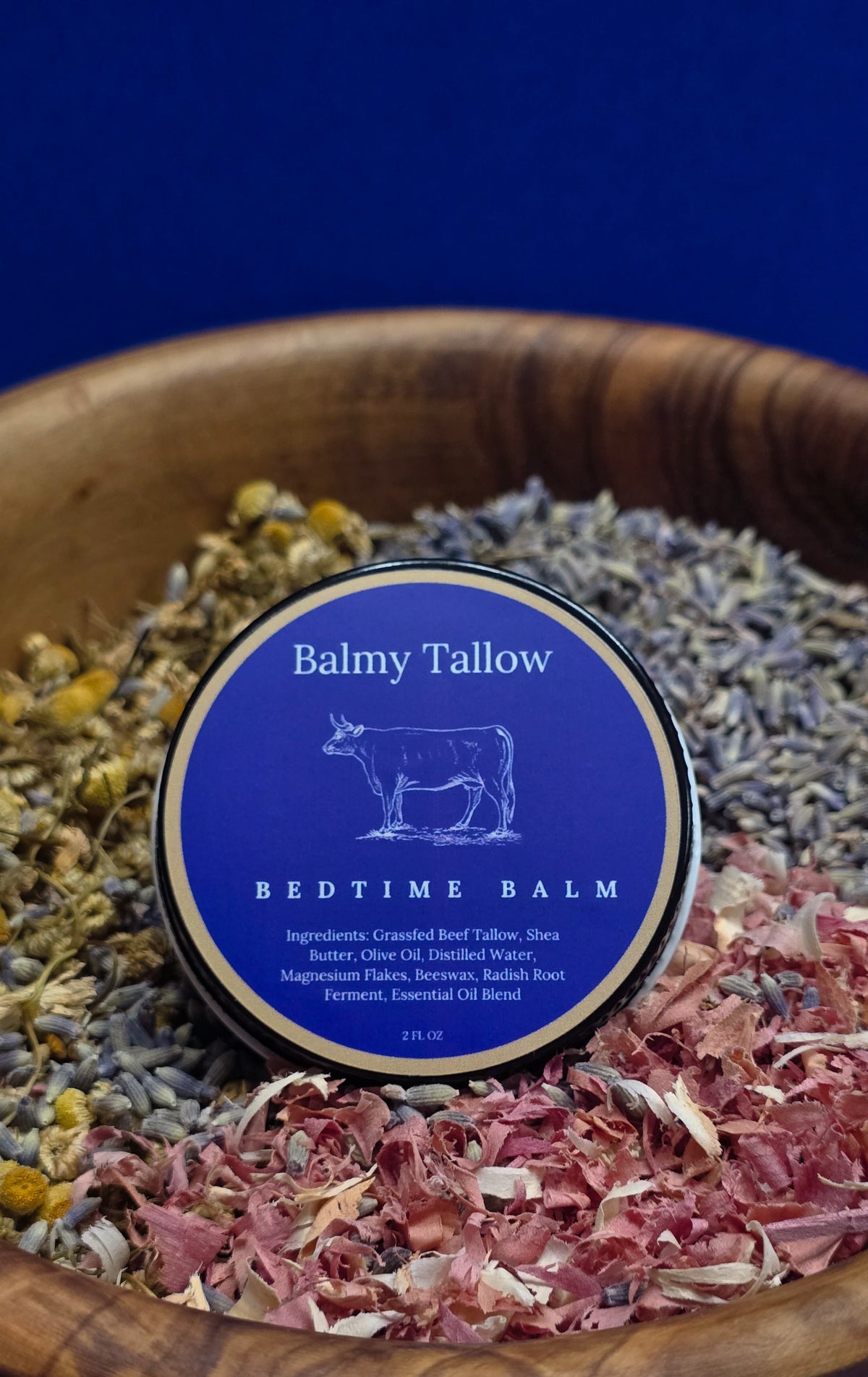 Balmy Tallow Bedtime Balm in an olive wood bowl with dried lavender, chamomile flowers, and cedarwood on a blue background