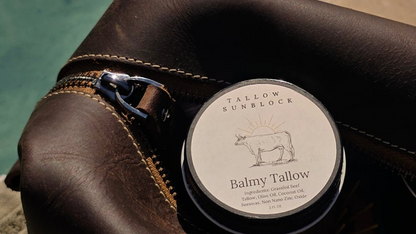 Close up of a round tin of Balmy Tallow Sunblock cream with a cow illustration on the label, resting on a brown leather pouch with a silver zipper pull.